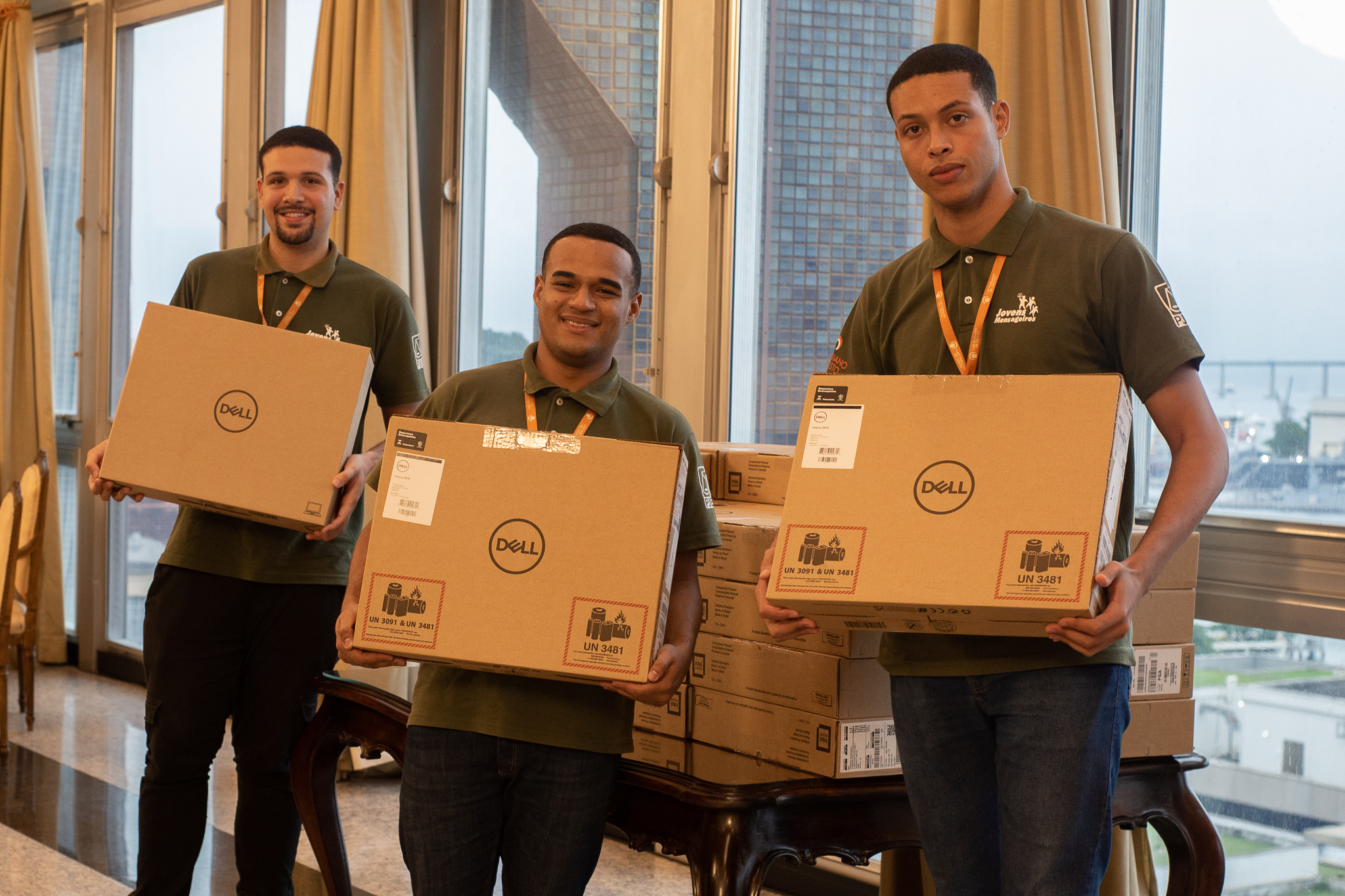 Foto posada de três participantes do projeto 'Jovens Mensageiros' com as caixas dos laptops nas mãos. 