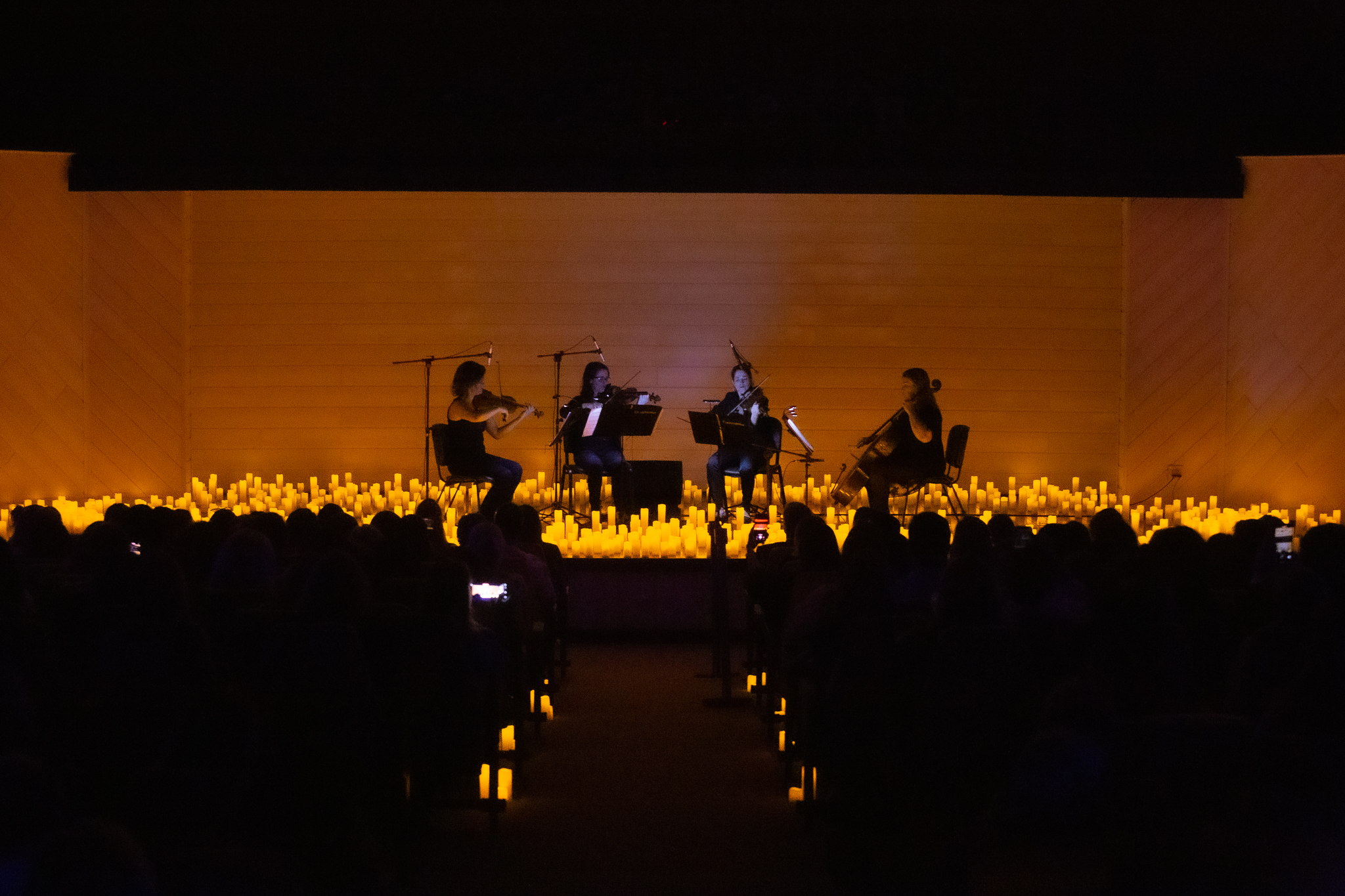 Imagem do auditório escuro, iluminado com velas de led no palco. No centro, quatro musicistas, sentadas, se apresentam com instrumentos de cordas