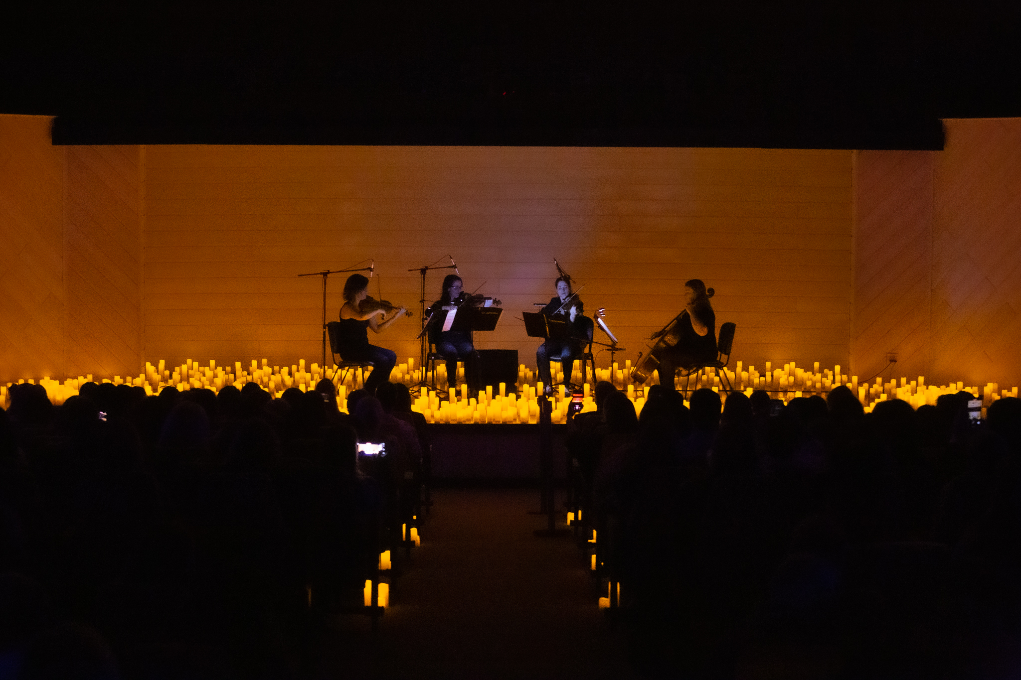 Imagem do auditório escuro, iluminado com velas de led no palco. No centro, quatro musicistas, sentadas, se apresentam com instrumentos de cordas 