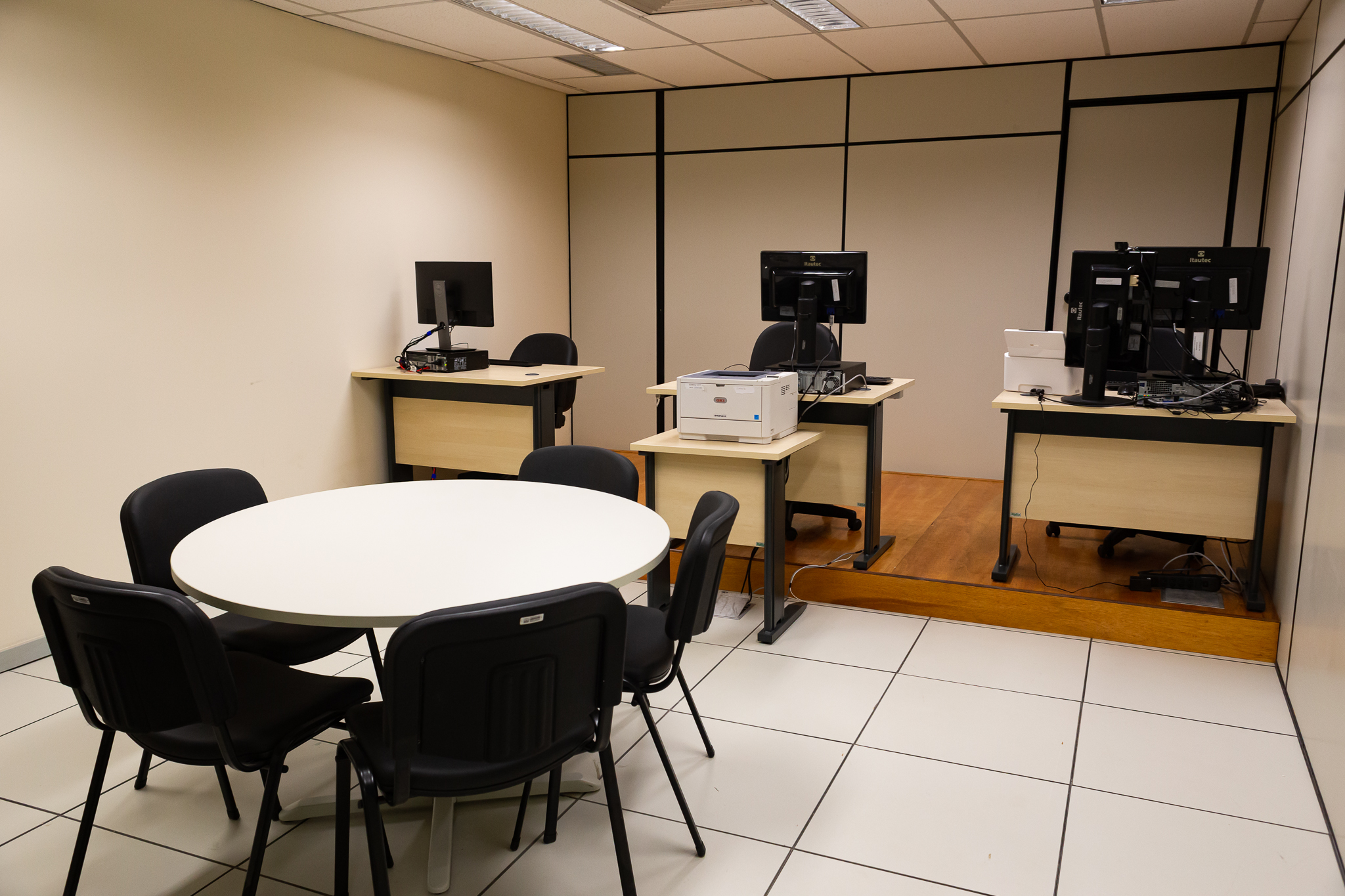 Imagem de uma das salas de audiência. Ao centro uma mesa redonda branca com cinco cadeiras pretas. Ao fundo, mais três mesas individuais com cadeira e computador 