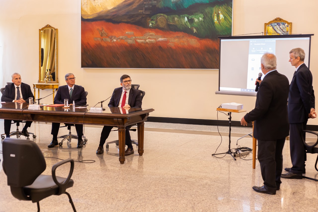 Dois desenvolvedores do aplicativo estão de pé apresentando o aplicativo, exibido num telão, aos integrantes da mesa - desembargador Marcos Chut, presidente do TJRJ, desembargador Henrique Figueira e presidente da OAB/RJ, Luciano Bandeira