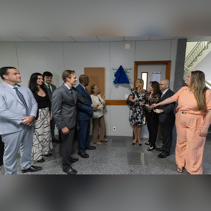 Grupo de pessoas de pé, em visão lateral, com placa no meio ao fundo, sendo descerrada