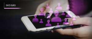 #ParaTodosVerem: Acima, no canto esquerdo, a palavra “DECISÃO”. A imagem mostra uma mão segurando um smartphone, com a tela voltada para cima. Sobre a tela do celular, há uma sobreposição gráfica de diversos ícones de pessoas em diferentes tamanhos e tons de roxo, representando indivíduos ou perfis. Esses ícones estão organizados de forma espalhada, criando a sensação de uma rede ou grupo. O fundo da imagem é escurecido nas bordas, com foco nas mãos e no celular, destacando o elemento central e a ideia de tomada de decisão baseada em dados, redes sociais ou participação coletiva via tecnologia.