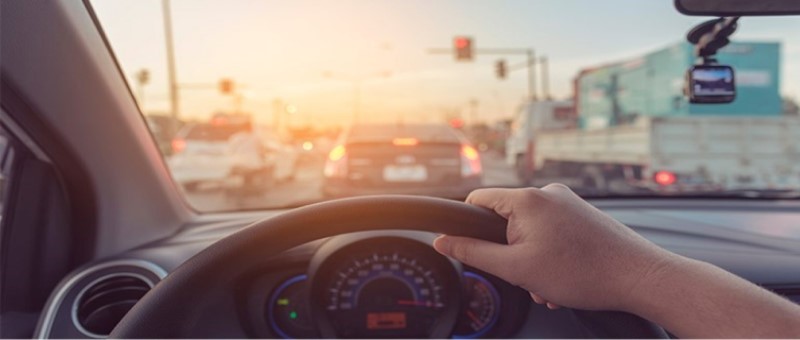 Imagem do trânsito intenso de uma rodovia ao fundo e, em primeiro plano,  visualizamos a mão do condutor ao volante e parte do painel do carro.