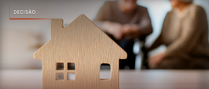 #ParaTodosVerem: Acima, no canto esquerdo, a palavra “DECISÃO”. Ao fundo, em imagem desfocada, um homem e uma mulher sentados conversando. À frente, sobre uma mesa, em imagem nítida, uma miniatura em madeira de uma casa.