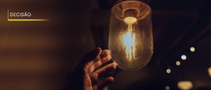 #ParaTodosVerem: Acima, à esquerda, o título “DECISÃO”. Numa fotografia com fundo bem escuro, mão de homem prestes a alcançar lâmpada queimada.