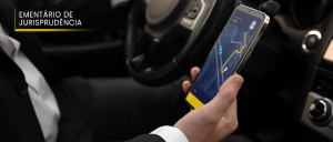 Imagem de uma pessoa ao volante de um carro, segurando em sua mão direita um celular com a imagem de um GPS em tela.