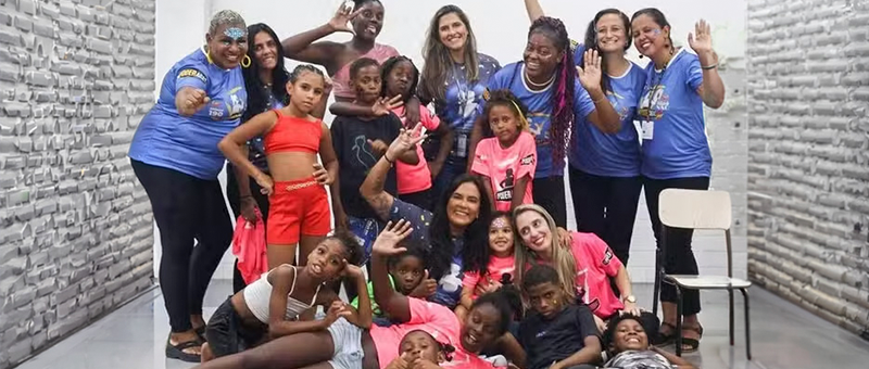 Foto mostra crianças e recreadores adultos sentados no chão em sala de paredes e piso brancos