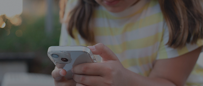 Criança segura um telefone celular com as duas mãos e olha para a tela, concentrada.