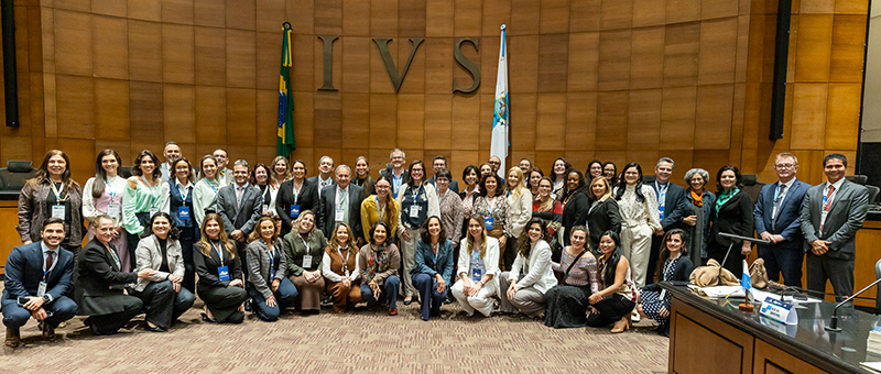 A imagem mostra uma fotografia de grupo realizada em um auditório institucional. Cerca de cinquenta pessoas posam para a foto, distribuídas entre fileiras em pé e agachadas na parte frontal. A maioria veste trajes sociais ou esporte fino, e muitos utilizam crachás de identificação pendurados no pescoço. Ao fundo, destaca-se uma parede revestida de madeira com a inscrição em letras grandes “IVS”, termo em latim associado ao conceito de Direito e Justiça. Também são visíveis as bandeiras do Brasil e do Estado do Rio de Janeiro, reforçando o caráter oficial do ambiente. À direita da imagem aparece uma mesa de autoridades, equipada com microfones e identificação institucional. 