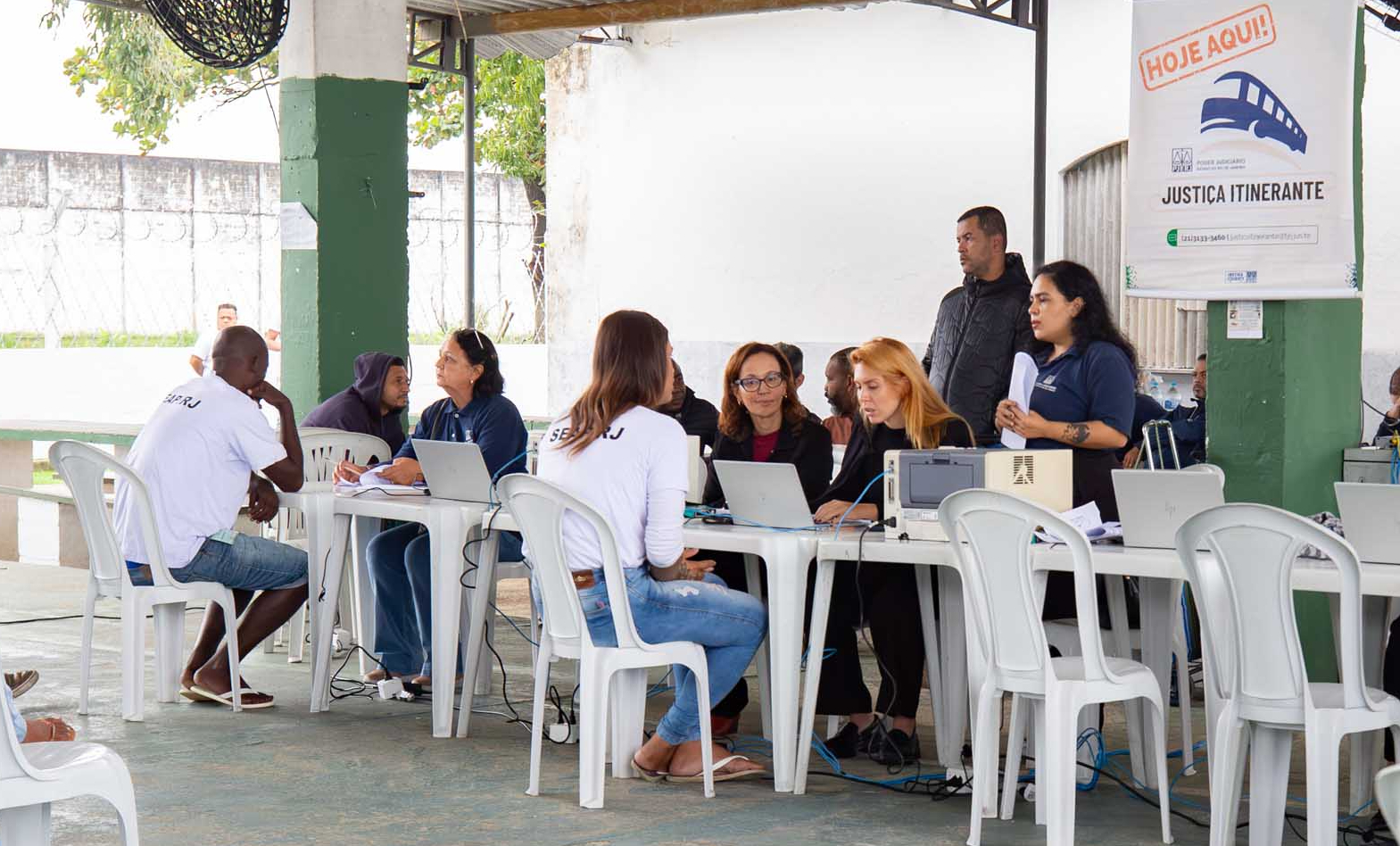 A imagem mostra uma ação da Justiça Itinerante realizada em um espaço coberto, com mesas organizadas para o atendimento ao público. Servidores e colaboradores do Poder Judiciário prestam orientações e realizam atendimentos individualizados, utilizando computadores e documentos durante o serviço. Em primeiro plano, um homem é atendido por uma servidora, enquanto outras equipes atendem diferentes pessoas ao longo das mesas. Ao fundo, há um banner com a identificação "Justiça Itinerante", acompanhado da inscrição "Hoje aqui!" e da logomarca do Poder Judiciário do Estado do Rio de Janeiro. O ambiente é simples e funcional, com cadeiras plásticas, equipamentos de informática e circulação de profissionais e usuários, evidenciando uma iniciativa voltada à ampliação do acesso à Justiça por meio de atendimento descentralizado.