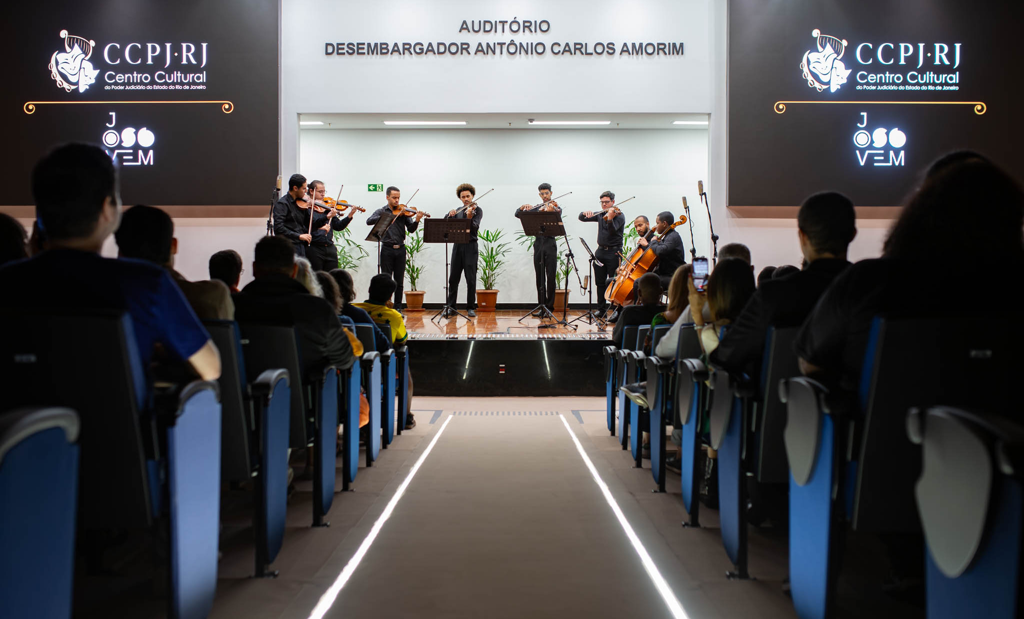 Em um auditório, um sexteto de música de câmara se apresenta em um palco diante de uma plateia. O grupo é formado por cinco músicos tocando violinos e um violoncelista, todos vestidos de preto e posicionados atrás de estantes para partituras. Ao fundo do palco, vasos com plantas ornamentam o ambiente. A fotografia foi registrada a partir do corredor central do auditório, destacando as fileiras de poltronas ocupadas pelo público, que acompanha atentamente a apresentação. À direita, uma pessoa registra o espetáculo com um telefone celular. Acima do palco, lê-se a inscrição "Auditório Desembargador Antônio Carlos Amorim". Nas duas telas laterais são exibidos o logotipo do Centro Cultural do Poder Judiciário do Estado do Rio de Janeiro (CCPJ-RJ) e a identidade visual do projeto Jovem, compondo o cenário do evento cultural.