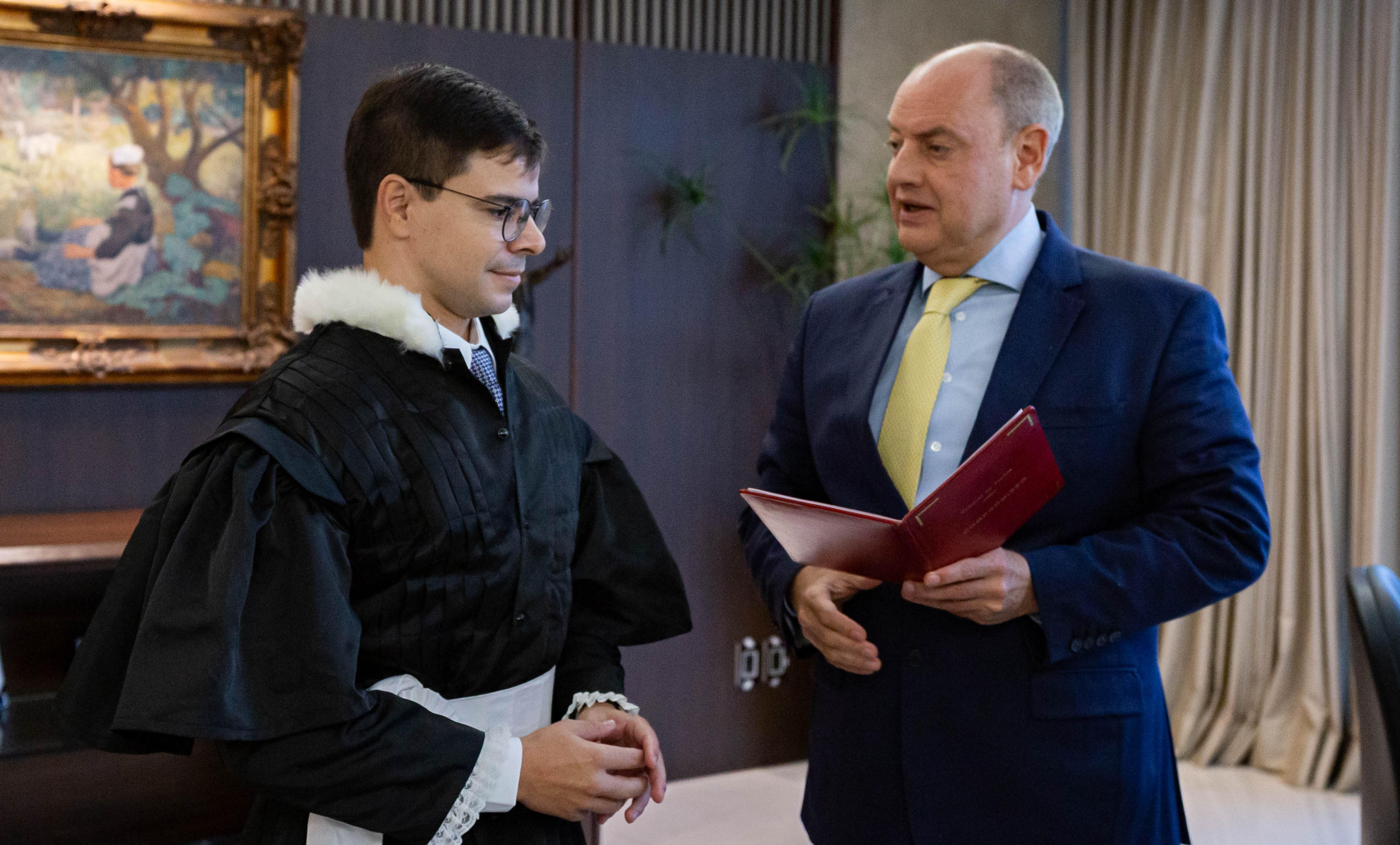 Foto mostra o juiz Felipe Lopes, homem branco, de cabelos escuros, vestindo toga preta com detalhes brancos na gola e punhos, próximo ao presidente do TJRJ, Des. Ricardo Couto, de terno azul marinho, camisa azul claro e gravata amarela, com livro de capa vermelha nas mãos, em ambiente fechado do gabinete da Presidência