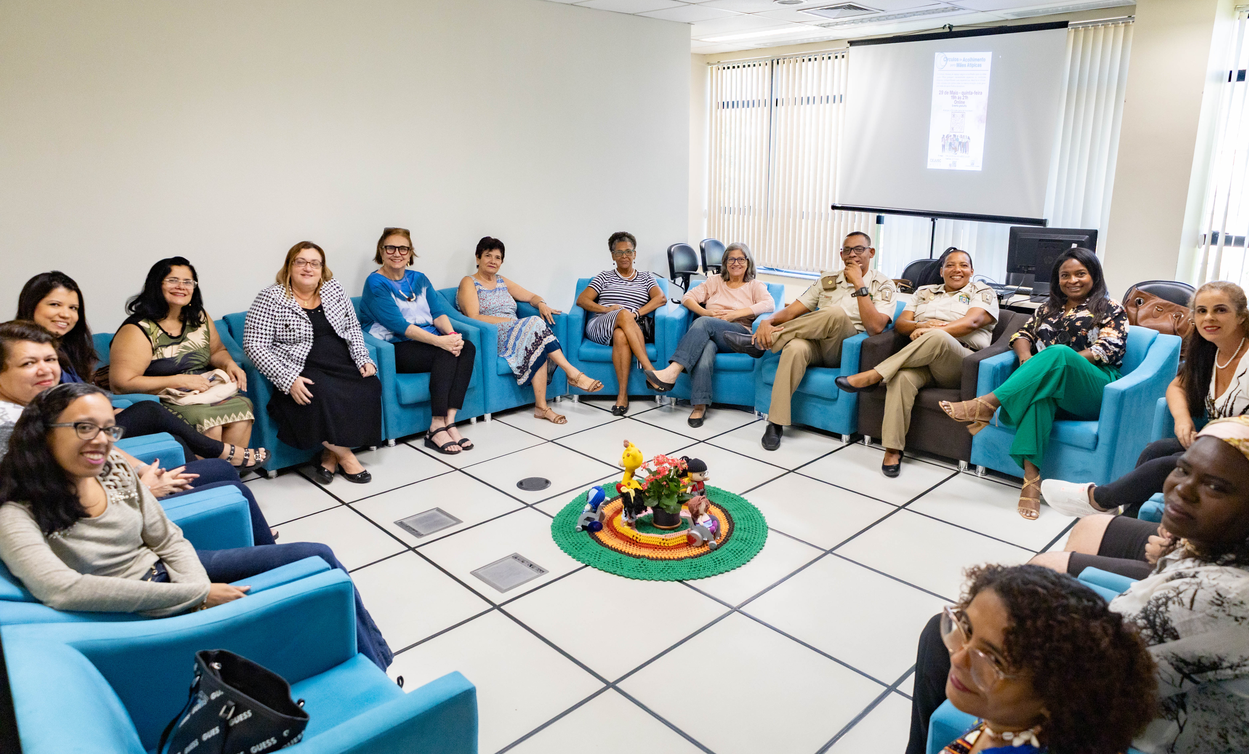 Foto mostra homens e mulheres sentados em poltronas azuis expostas de maneira circular em sala com pintura e piso claros. Ao centro, um pequeno tapete redondo colorido tem sobre ele um vaso de flor e alguns brinquedos