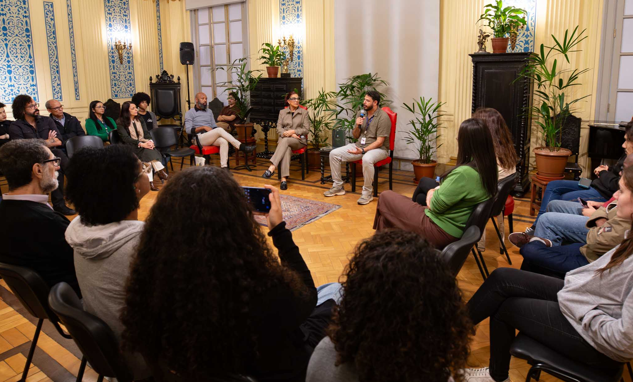 A imagem mostra uma roda de conversa realizada em um salão histórico, com participantes sentados em círculo para acompanhar o debate. No centro, um homem segura um microfone enquanto fala ao público, ao lado de uma mulher também sentada, que acompanha a discussão. Os participantes, distribuídos em cadeiras ao redor dos debatedores, ouvem atentamente a apresentação. Em primeiro plano, uma pessoa registra o encontro com um celular. O ambiente é amplo e elegante, com piso de madeira, paredes ornamentadas com detalhes em azul e branco, grandes colunas, luminárias clássicas e diversas plantas distribuídas pelo espaço, conferindo um clima acolhedor. Ao fundo, móveis de época e amplas portas reforçam o caráter histórico do local.