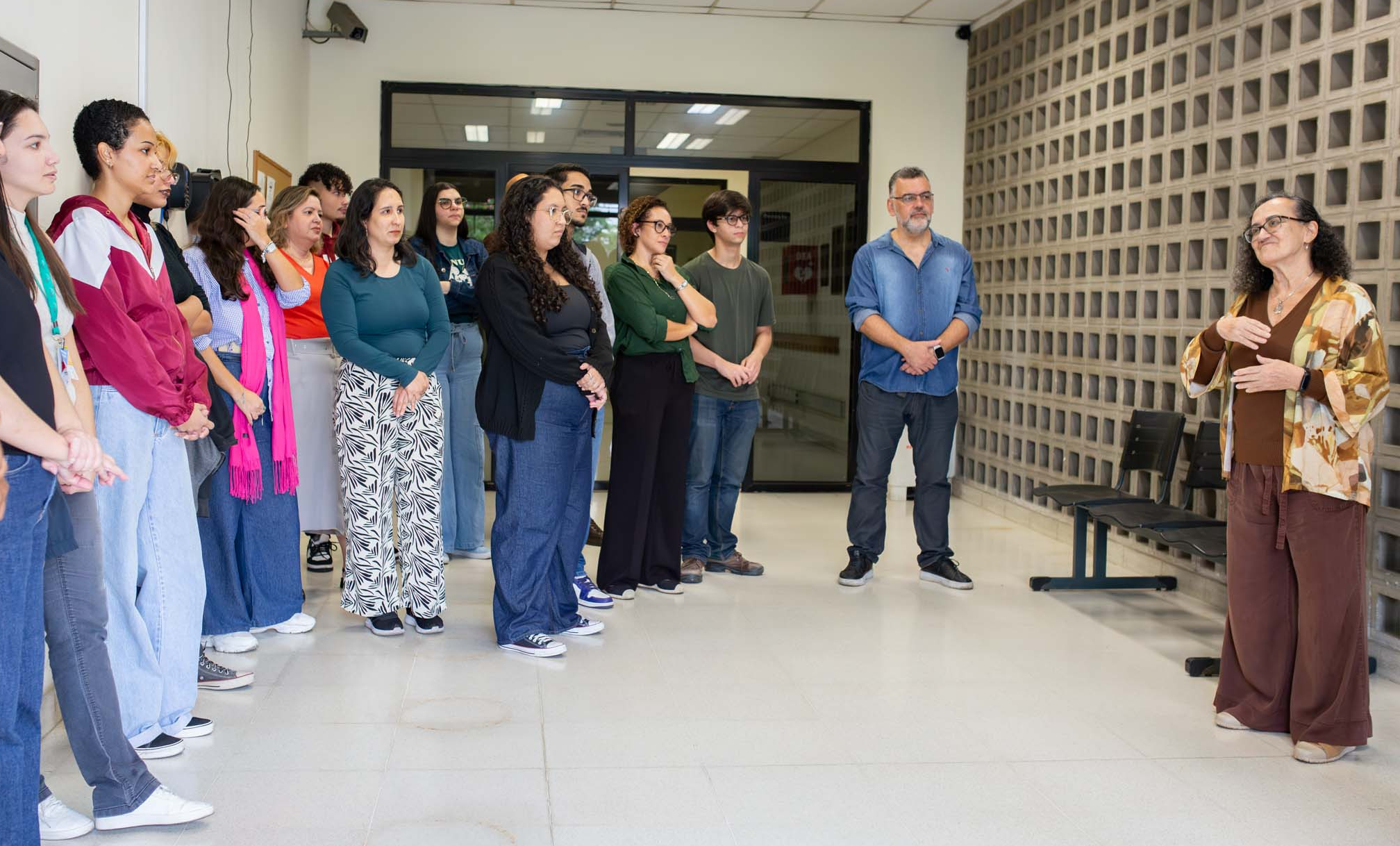 A imagem mostra um grupo de visitantes reunido em um espaço interno durante uma atividade de visita guiada. Os participantes estão posicionados em semicírculo, atentos à fala de uma mulher que conduz a apresentação. Ela gesticula enquanto se dirige ao grupo, que acompanha a explicação com atenção. O grupo é composto por jovens e adultos, vestidos de maneira casual, distribuídos ao longo de um amplo corredor iluminado. Ao fundo, há portas de vidro e uma parede com elementos vazados em concreto, que conferem identidade arquitetônica ao ambiente. Próximo à mediadora, algumas cadeiras estão alinhadas junto à parede.