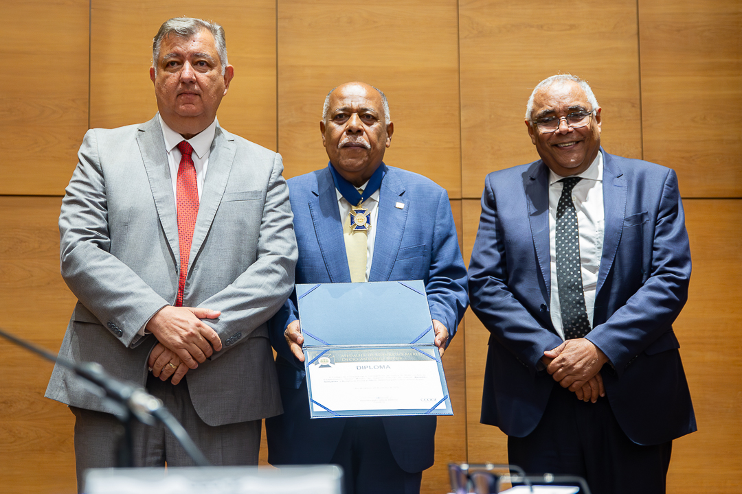 A imagem mostra três homens em pé, lado a lado, em um ambiente interno com parede revestida de painéis de madeira clara ao fundo. O homem ao centro veste terno azul e gravata clara. Ele usa no pescoço uma medalha presa por uma fita azul e segura com as duas mãos um diploma aberto, exibindo o documento para a câmera. Sua expressão é séria e solene. À esquerda, há um homem de terno cinza claro e gravata vermelha. Ele está com as mãos unidas à frente do corpo e mantém uma expressão mais neutra, olhando diretamente para a câmera. À direita, outro homem veste terno azul escuro, camisa branca e gravata escura com pequenos pontos claros. Ele está com as mãos cruzadas à frente do corpo e apresenta um leve sorriso.