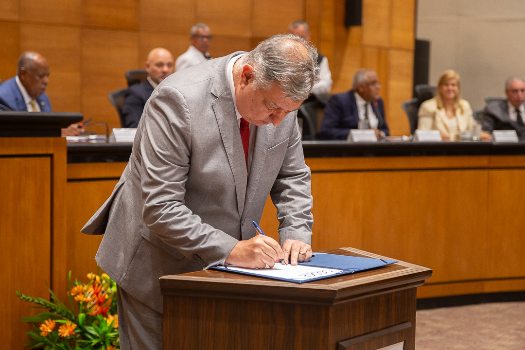 A imagem mostra o desembargador Cláudio Brandão de Oliveira vestindo terno cinza e gravata vermelha assinando um documento sobre um púlpito de madeira. Ele está inclinado para frente, concentrado, segurando uma caneta enquanto escreve em uma pasta azul aberta. O local é o Tribunal Pleno do TJRJ que conta com revestimento de madeira nas paredes. Ao fundo, há várias pessoas sentadas em uma bancada elevada, também vestidas formalmente, observando o momento. Na parte inferior à esquerda da imagem, há um arranjo de flores com tons de laranja e verde.