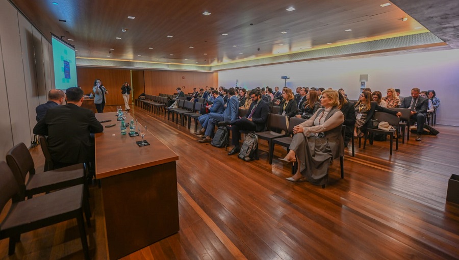 A imagem mostra uma sala com cadeiras enfileiradas e uma mulher fazendo uma apresentação ao lado de um telão em frente a plateia. 