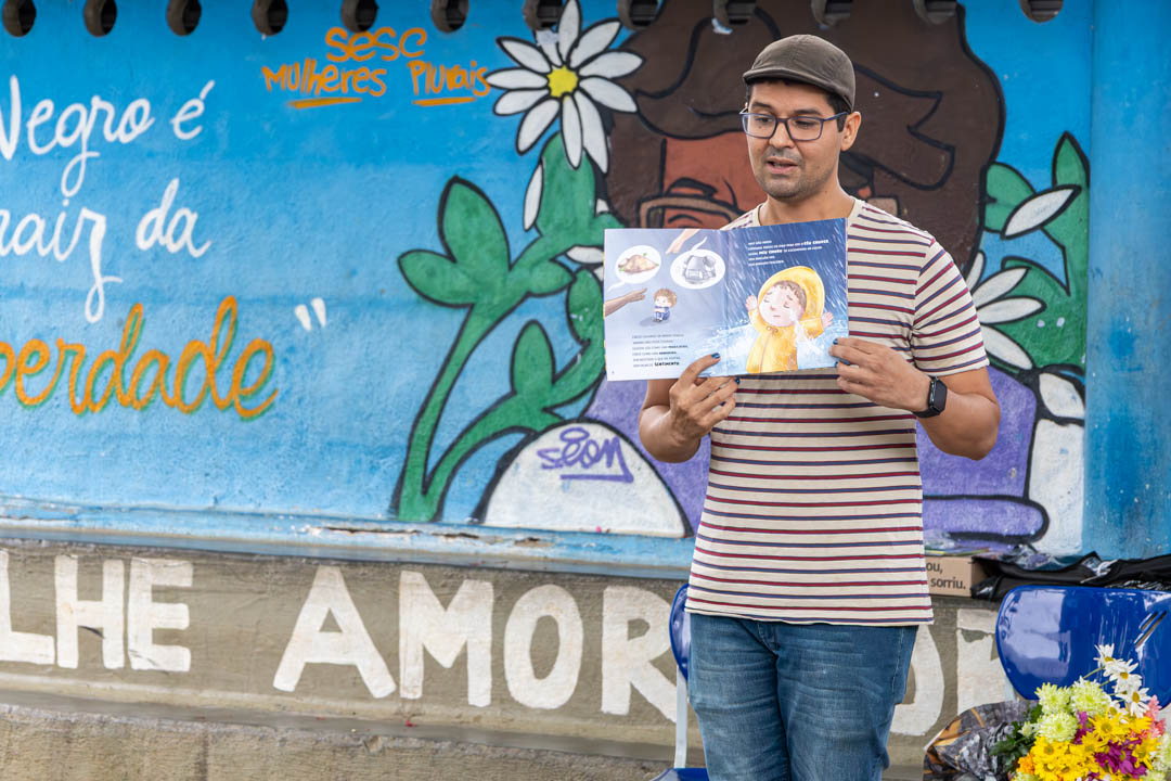 A imagem apresenta um homem em pé de blusa listrada e óculos mostrando um livro aberto