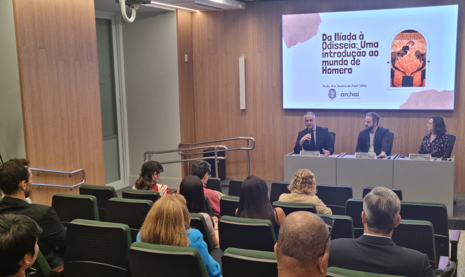 A imagem mostra um auditório cheio com três debatedores na mesa principal (dois homens e uma mulher)