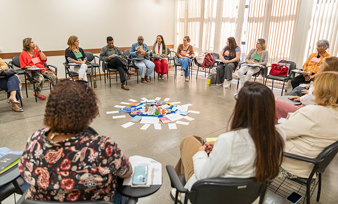 A imagem mostra um grupo de pessoas sentadas em círculo durante uma apresentação em uma sala de aula. 