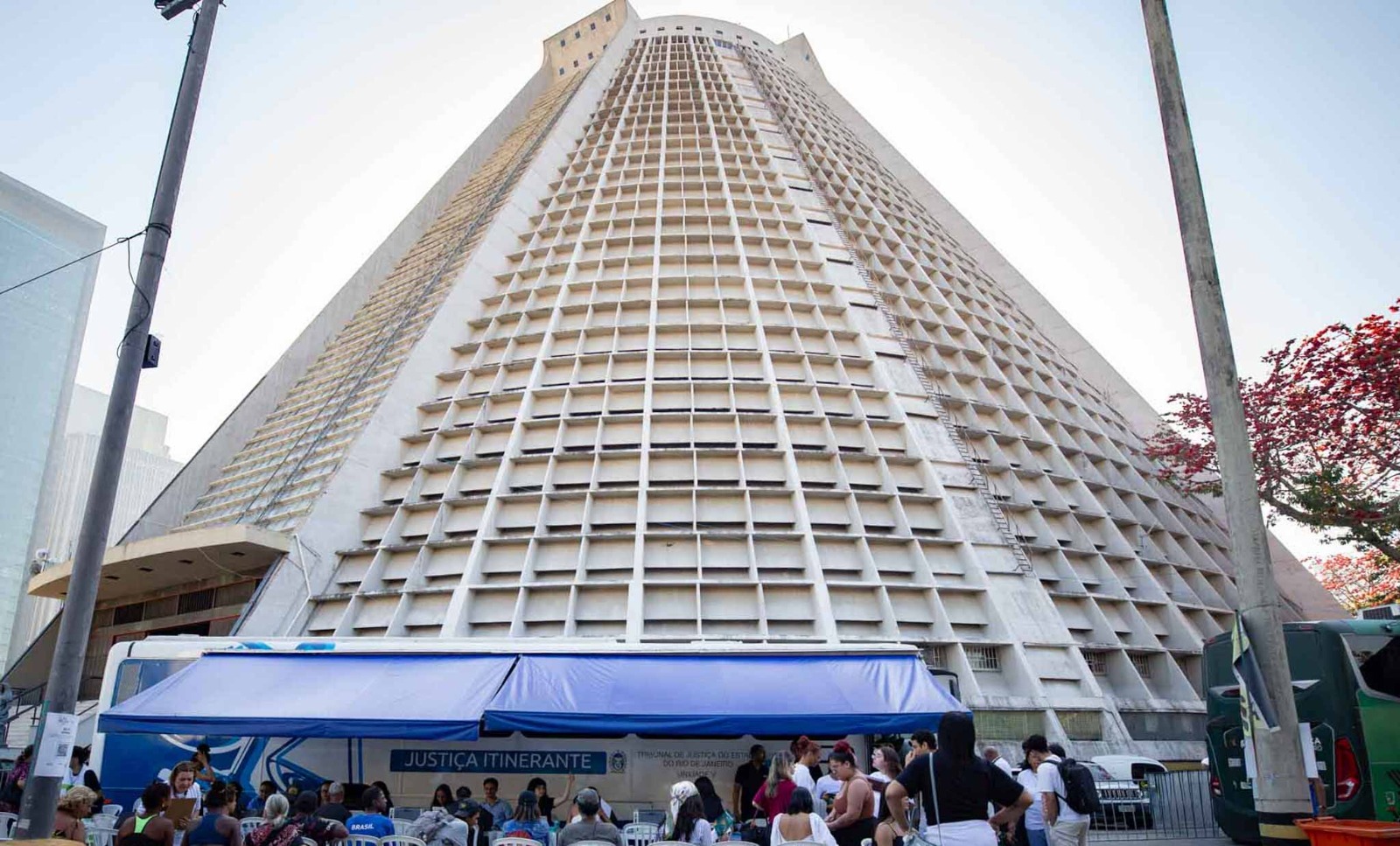 A fotografia mostra a fachada do edifício da Catedral Metropolitana de São Sebastião, registrada de baixo para cima, destacando sua arquitetura imponente e a estrutura em concreto com brises geométricos. Em primeiro plano, está montada uma tenda azul da Justiça Itinerante, onde servidores prestam atendimento ao público. Em frente à tenda, dezenas de pessoas aguardam atendimento, algumas sentadas em cadeiras brancas organizadas em fileiras e outras em pé, formando pequenos grupos. Sobre a estrutura da tenda, é possível ler a identificação "Justiça Itinerante" e o nome do Tribunal de Justiça do Estado do Rio de Janeiro.