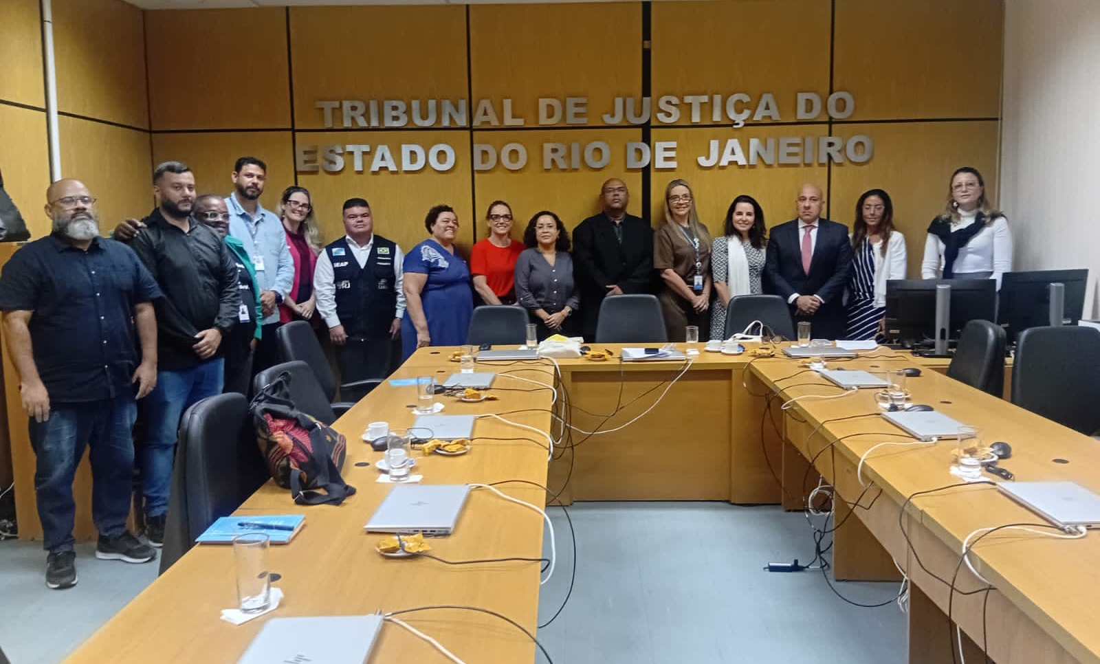 Sala com uma mesa de madeira em formato de U com 15 pessoas de pé atras, escrito Tribunal de Justiça do Estado do Rio de Janeiro em letras metálicas na parede ao fundo