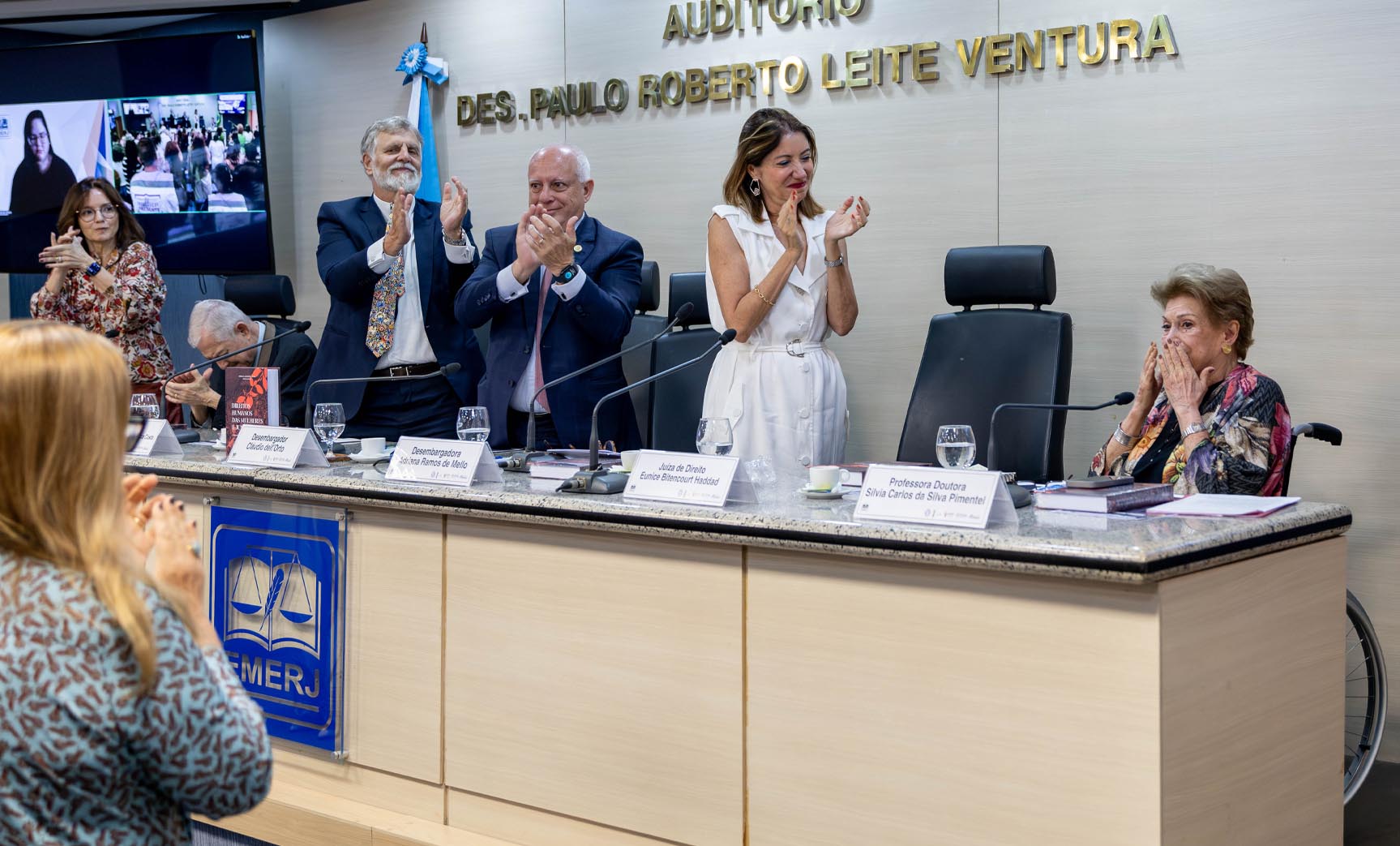 A imagem mostra pessoas em pé em um auditório aplaudindo a homenageada, sentada à direita com as mãos no rosto emocionada
