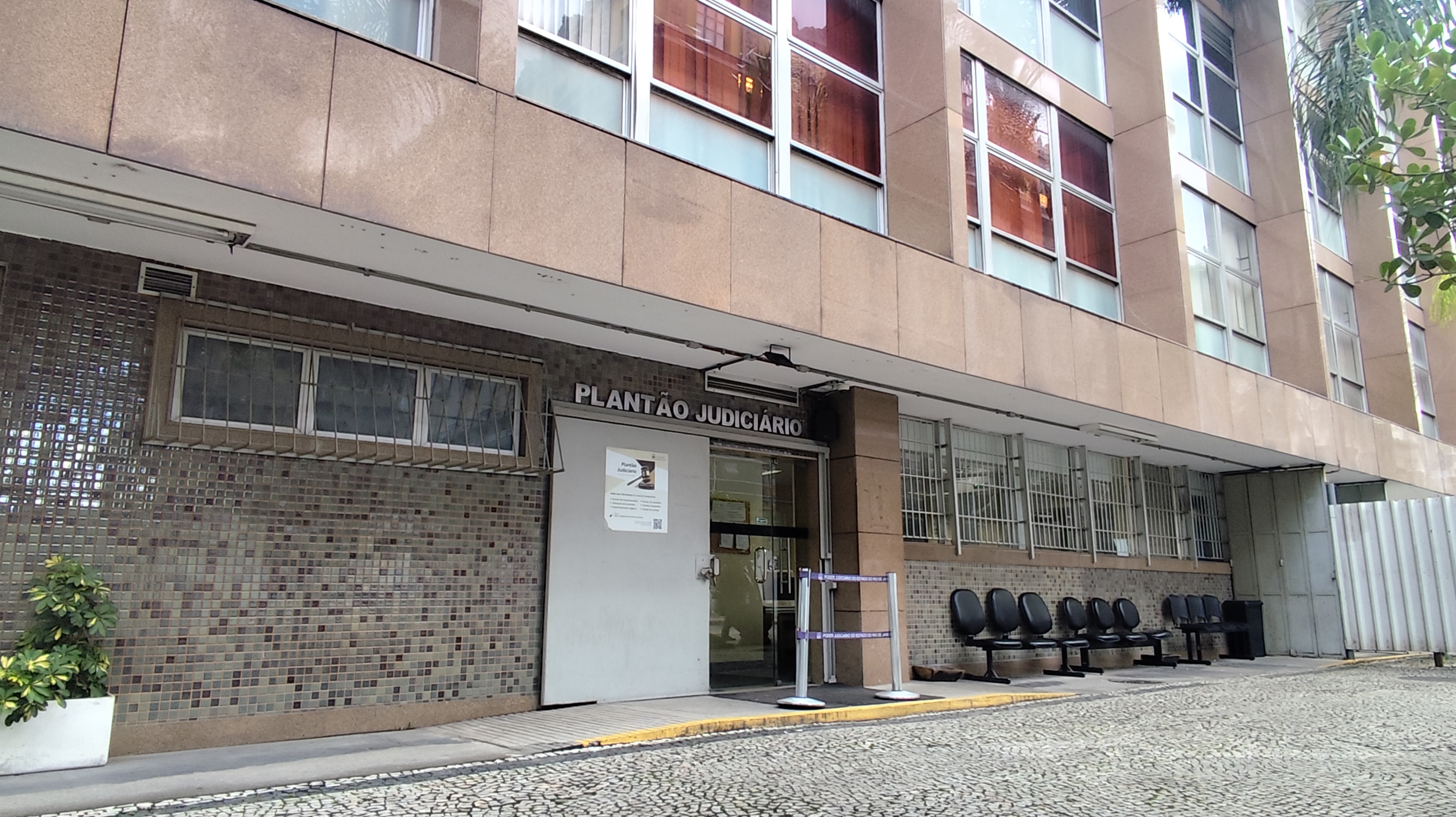 A imagem mostra a fachada de um edifício onde funciona o Plantão Judiciário. O prédio possui revestimento em tons de bege e marrom, com janelas amplas distribuídas pelos andares superiores. Acima da entrada principal, há uma placa com a inscrição “PLANTÃO JUDICIÁRIO”. Na parte térrea, a fachada é revestida com pequenos azulejos em tons de cinza e marrom. As janelas possuem grades de proteção metálicas. Ao lado da porta de entrada, há um cartaz afixado na parede e uma pequena área delimitada por fitas organizadoras. Em frente ao prédio, há uma fileira de cadeiras pretas para espera. O acesso ao edifício é feito por uma área pavimentada com pedras portuguesas em padrão ondulado, típica das calçadas do Rio de Janeiro. Nas extremidades da imagem, é possível observar vegetação, incluindo um vaso com planta à esquerda e árvores à direita.