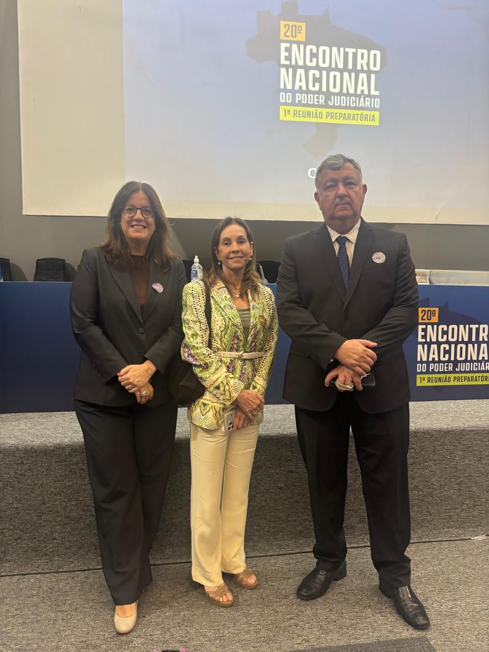 Foto com duas magistradas à esquerda e o corregedor à direita. No fundo, há um telão com imagem "20º Encontro Nacional do Poder Judiciário - 1ª Reunião Preparatória"