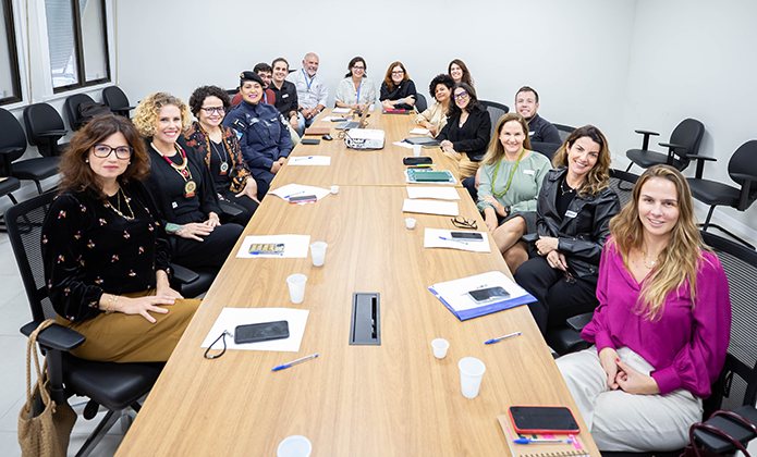 A imagem mostra uma sala de reunião com uma mesa retangular no centro e cerca de 20 pessoas ao redor dela