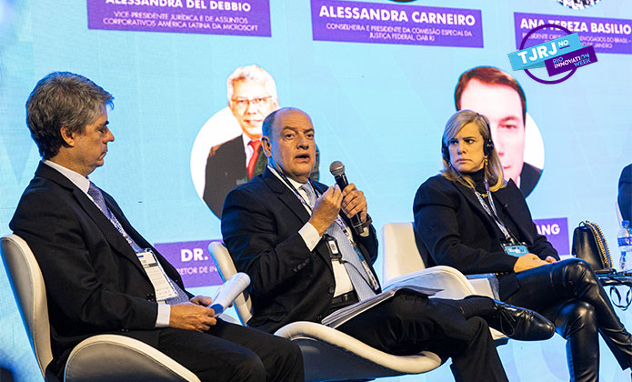 A imagem mostra três pessoas - dois homens, um deles é o presidente do TJRJ ao centro, e uma mulher - sentados em cadeiras no palco durante um debate com um telão ao fundo.