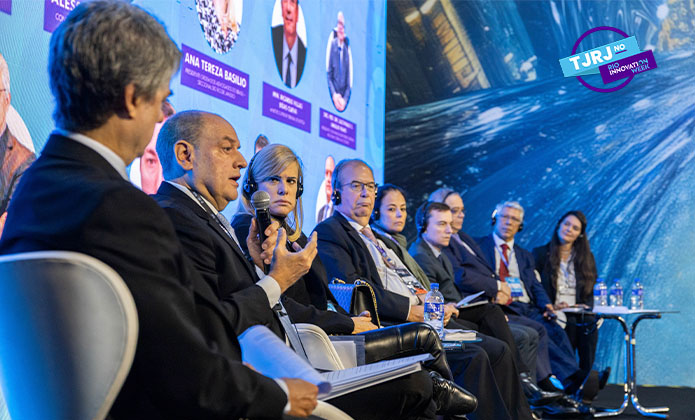 A imagem mostra nove pessoas sentadas em cadeiras no palco participando de um debate. O fundo é azul e tem um telão com fotos e nomes dos participantes do evento. 