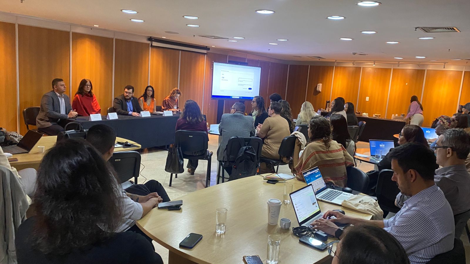 A foto mostra cerca de 20 pessoas espalhadas em mesas com laptops em uma sala e uma mesa principal na frente com cinco pessoas apresentando uma aula. Há um telão ao lado mostrando informações da apresentação. 