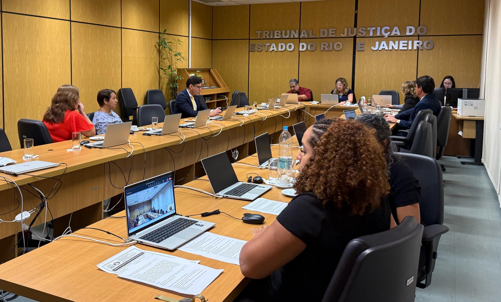 A imagem mostra uma reunião em andamento em uma sala do Tribunal de Justiça do Estado do Rio de Janeiro. Várias pessoas estão sentadas ao redor de uma mesa retangular, equipadas com notebooks, papéis e copos de água. A sala tem painéis de madeira nas paredes e um letreiro prateado com o nome da instituição. 