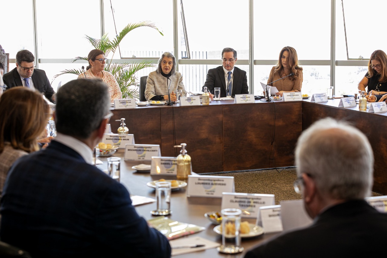 A foto mostra uma sala de reuniões com mesa em formato de "U" e os participantes sentados ao redor. 