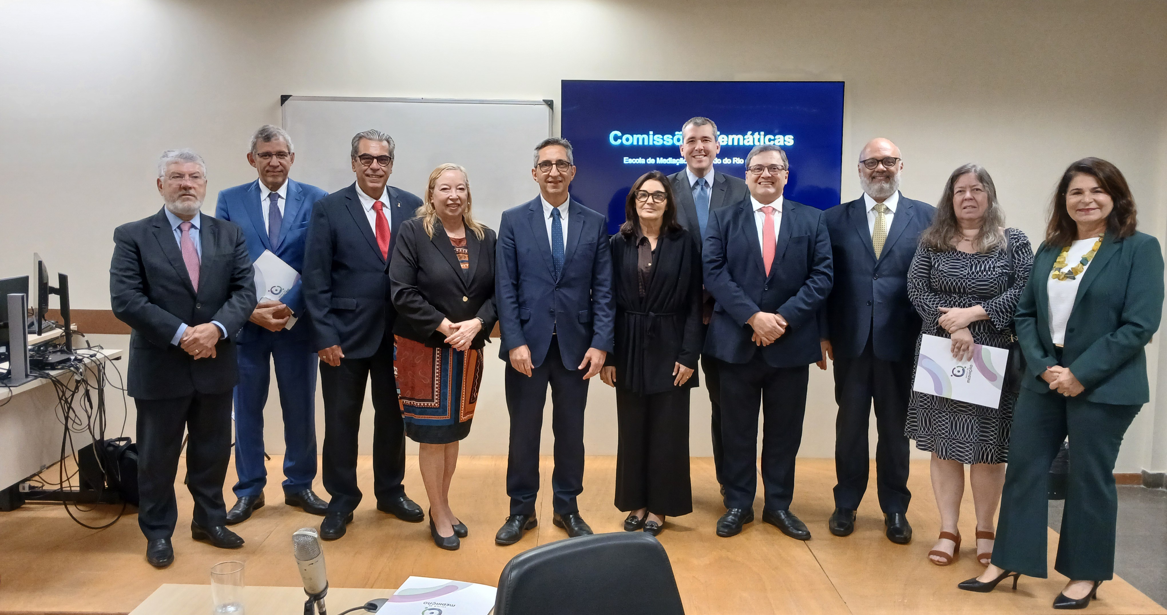 A imagem mostra um grupo de integrantes das Comissões Temáticas reunidos para uma fotografia institucional em uma sala de reuniões da Escola de Mediação do Tribunal de Justiça do Estado do Rio de Janeiro (EMEDI). Ao todo, onze pessoas — homens e mulheres — estão alinhadas lado a lado, de frente para a câmera. A maioria veste trajes formais, como ternos escuros, gravatas e conjuntos sociais, característicos de eventos institucionais do meio jurídico. Alguns seguram pastas ou documentos. Ao fundo, um telão exibe a inscrição “Comissões Temáticas – Escola de Mediação do Tribunal de Justiça do Estado do Rio de Janeiro”, indicando que o registro foi feito durante um encontro relacionado às atividades da Emedi. O ambiente possui paredes claras e piso de madeira, com estrutura típica de sala de reuniões ou auditório.