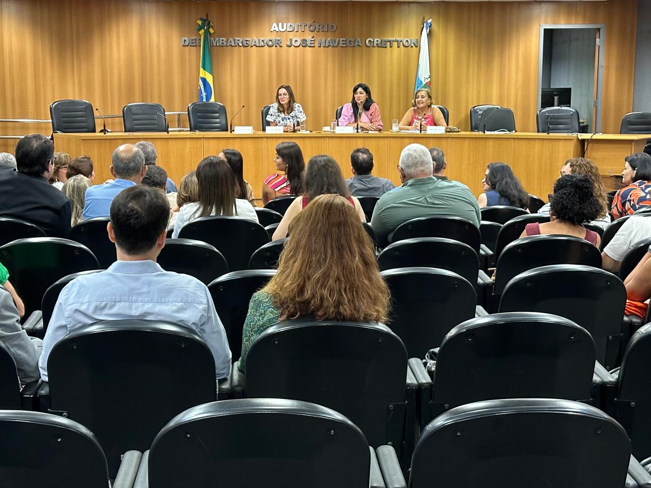 A imagem mostra um auditório com pessoas assistindo a uma reunião. Na parte da frente, há uma mesa com três mulheres sentadas, falando com a plateia. No fundo, na parede de madeira, está escrito “AUDITÓRIO DESEMBARGADOR JOSÉ NAVEGA CRETTON”. Também é possível ver bandeiras ao fundo, sendo uma do Brasil e outra do Estado do Rio de Janeiro. A plateia é composta por homens e mulheres vestidos de maneira formal, e algumas pessoas estão interagindo entre si. 