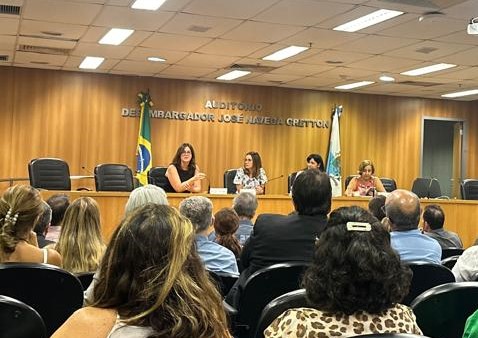 A imagem mostra um auditório onde ocorre uma reunião institucional. Na parte da frente, há uma mesa com quatro mulheres sentadas, participando de um painel de discussão. Uma delas está falando enquanto as outras ouvem. O fundo da sala possui um painel de madeira com o nome “AUDITÓRIO DESEMBARGADOR JOSÉ NAVEGA CRETTON”, além de bandeiras, incluindo a do Brasil. A plateia é composta por diversas pessoas, algumas vestidas formalmente e outras de maneira mais casual, que estão atentas ao evento. O ambiente tem iluminação artificial e cortinas fechadas ao fundo.