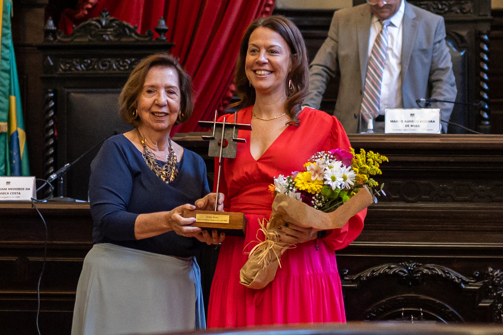 ​ A cerimônia foi presidida pelo diretor-geral da Emerj, desembargador Marco Aurélio Bezerra de Melo, e teve como madrinhas as magistradas Myriam Medeiros da Fonseca Costa, filha da jurista Romy Martins Medeiros da Fonseca; e a desembargadora Cristina Tereza Gaulia, presidente do Fórum Permanente de Diálogos da Lei com o Inconsciente e do Fórum Permanente de Estudos Constitucionais, Administrativos e de Políticas Públicas Professor Miguel Lanzellotti Baldez.     As seis mulheres homenageadas na edição de 2024 foram a tabeliã de notas do Estado de São Paulo Carla Watanabe; a juíza Eunice Bitencourt Haddad, presidente da Associação dos Magistrados do Estado do Rio de Janeiro (Amaerj); a atriz Fernanda Montenegro; a historiadora Lilia Moritz Schwarcz; a ministra do Superior Tribunal de Justiça (STJ) Regina Helena Costa e a desembargadora Renata Machado Cotta.                                                 Escola da Magistratura do Rio homenageou as mulheres que se destacaram na magistratura, política e arte     Tabeliã Carla Watanabe  A doutoranda e mestre em Direito Constitucional pelo Instituto Brasileiro de Ensino Desenvolvimento e Pesquisa (IDP) Carla Watanabe se destacou na luta pelos direitos de pessoas transexuais e foi a primeira tabeliã de notas trans no país. “Estar aqui é algo muito forte e de muita responsabilidade para mim porque carrego aqueles que, assim como eu, sofreram discriminação. Esse prêmio é um reconhecimento da nossa luta”, pontuou.   Juíza Eunice Bitencourt Haddad  A juíza Eunice Bitencourt Haddad, que está em seu segundo biênio como presidente da Amaerj, já fez parte do Conselho Deliberativo e Fiscal em 2020 e 2021, e foi juíza auxiliar da Presidência do Tribunal de Justiça do Rio em 2020. “Devemos ser inspiração para quem estiver ao nosso redor, construindo uma corrente do bem e de igualdade para vivermos em um mundo melhor”, afirmou.     A presidente da Amaerj Eunice Bitencourt Haddad recebeu o prêmio das mães da desembargadora aposentada Myriam Medeiros da Fonseca Costa   ​