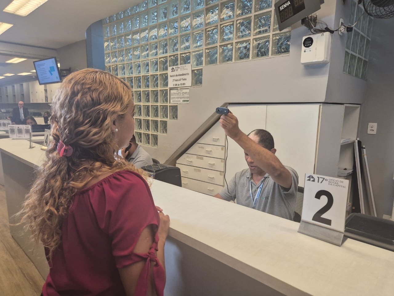 A imagem mostra uma cena em um cartório. No primeiro plano, uma mulher está de pé em frente a um balcão, vista de costas e de perfil. Ela tem cabelos longos e cacheados, presos parcialmente, e veste uma blusa de cor vinho. Do outro lado do balcão, um atendente está sentado e segura uma câmera apontada em direção à mulher, como se estivesse registrando uma foto ou coletando dados. Sobre o balcão há um suporte com o número 2, indicando o guichê de atendimento.