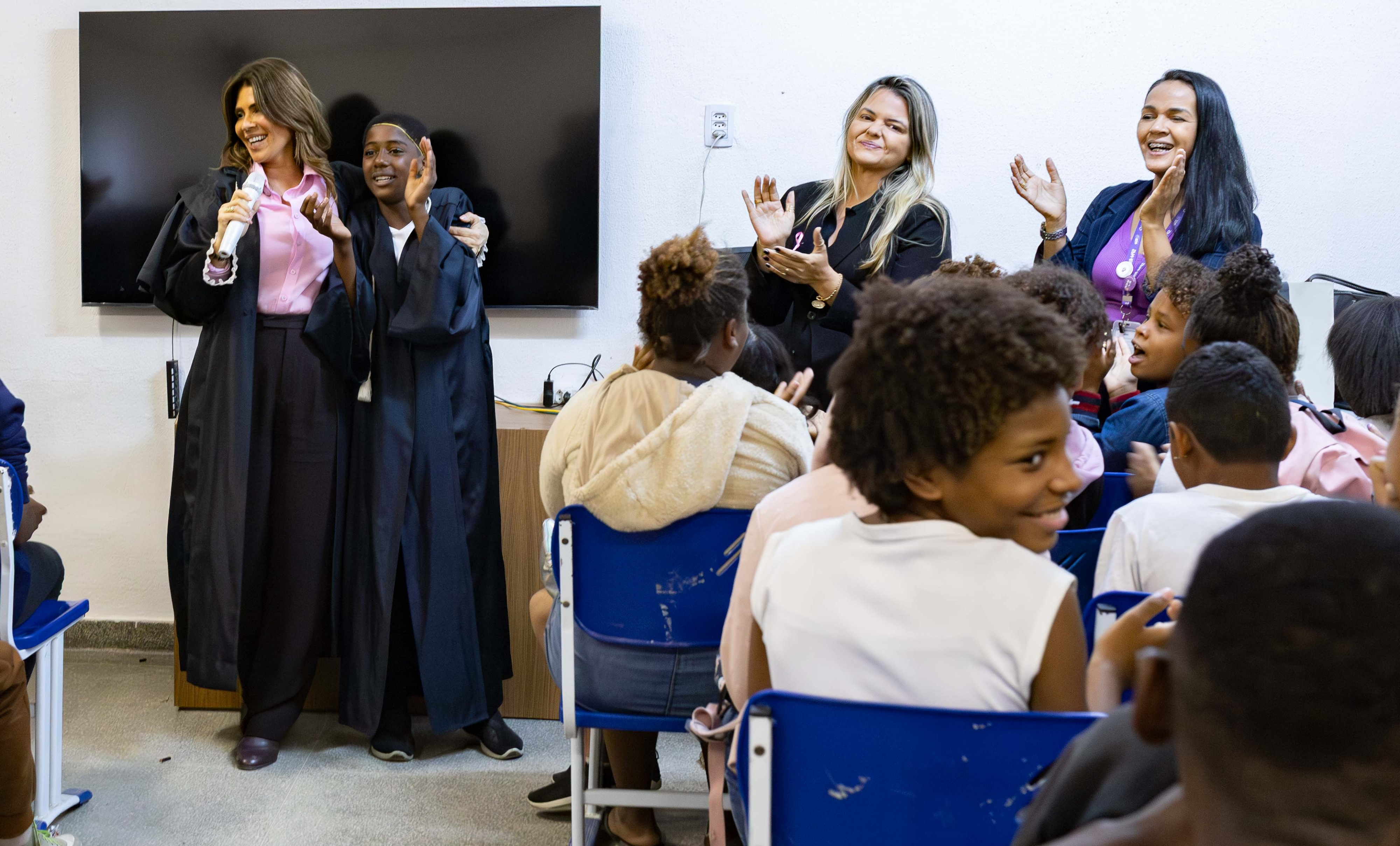 Imagem da juíza Daniella Alvarez Prado abraçando o aluno Riquelme Rodrigues. De costas alunos aplaudem ação 