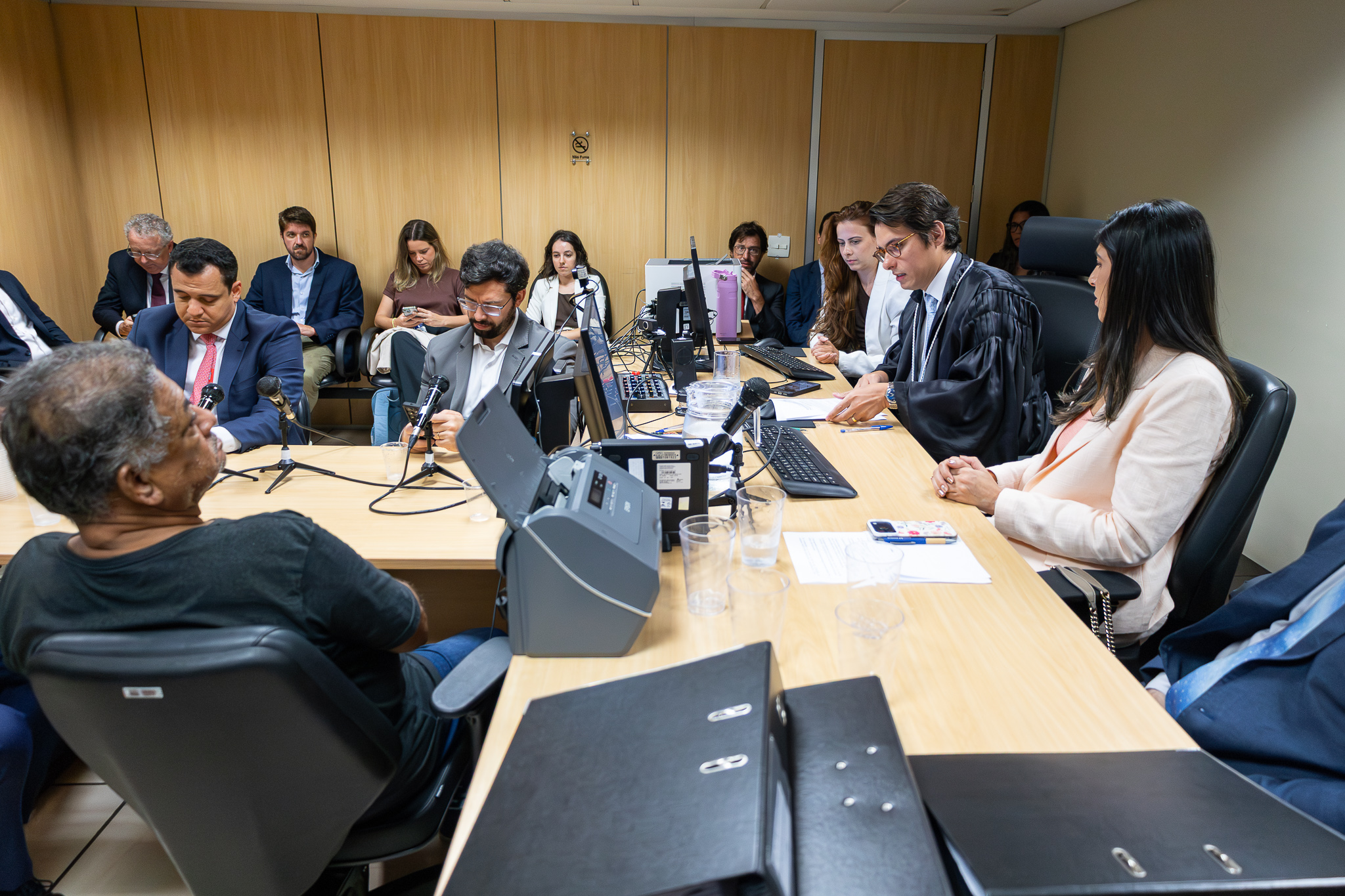 A imagem mostra uma sala de audiência com várias pessoas sentadas ao redor de uma mesa retangular de madeira clara. O ambiente é fechado, com paredes revestidas em painel de madeira e iluminação artificial no teto. No lado direito da mesa, um homem veste toga preta, indicando tratar-se de um magistrado. Ele está lendo ou consultando documentos posicionados diante de um computador. Ao seu lado, uma mulher com blazer claro acompanha a sessão com postura atenta. À esquerda da mesa, há outros participantes, incluindo advogados e representantes, vestidos com ternos e trajes sociais. Um homem segura um microfone de mesa, e há outros microfones posicionados sobre a superfície, sugerindo que a sessão está sendo gravada ou transmitida. Também são visíveis computadores, cabos, copos plásticos com água, documentos e equipamentos eletrônicos.