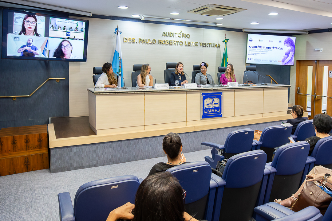 A imagem mostra evento realizado no Auditório Desembargador Paulo Roberto Leite Ventura, na sede da Escola da Magistratura do Estado do Rio de Janeiro (EMERJ). Na parte frontal do auditório, cinco mulheres estão sentadas atrás de uma mesa de debates, participando de uma mesa-redonda. A mesa é composta pela superintendente Taiza Moreno, a juíza Michela Vechi, a desembargadora Adriana Ramos, a conselheira Fátima Cidade e a promotora Isabela Jourdan palestraram na segunda mesa. Cada uma delas tem à frente um microfone, copos de água e placas de identificação com seus nomes e funções. Ao centro da mesa aparece o logotipo da EMERJ. Ao fundo, na parede, está o nome do auditório em letras douradas, além das bandeiras do Brasil e do Estado do Rio de Janeiro. À esquerda do palco, um telão exibe participantes conectadas por videoconferência e a presença de uma intérprete de Libras. À direita, outro telão apresenta o cartaz do evento, cujo tema é “A violência obstétrica: uma grave violação aos direitos das mulheres”. Na plateia, diversas pessoas estão sentadas em cadeiras azuis, acompanhando a discussão.