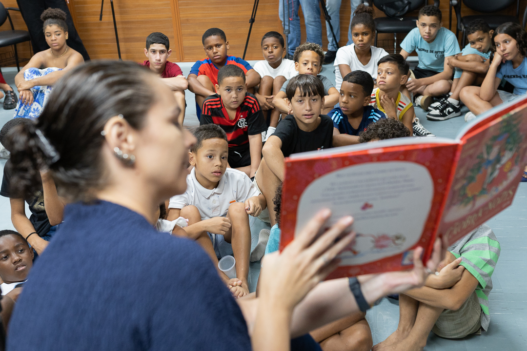 Imagem de grupo de crianças de frente observando a assistente de direção do CCPJ Roberta Araújo durante contação de história