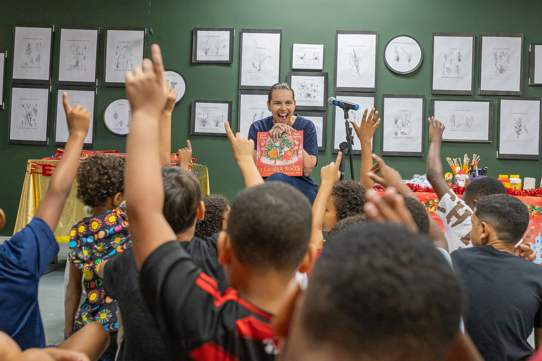 Imagem de crianças de costas com os braços para o alto fazendo sinal de presente e ao fundo, assistente de direção do CCPJ Roberta Araújo, de frente para as crianças