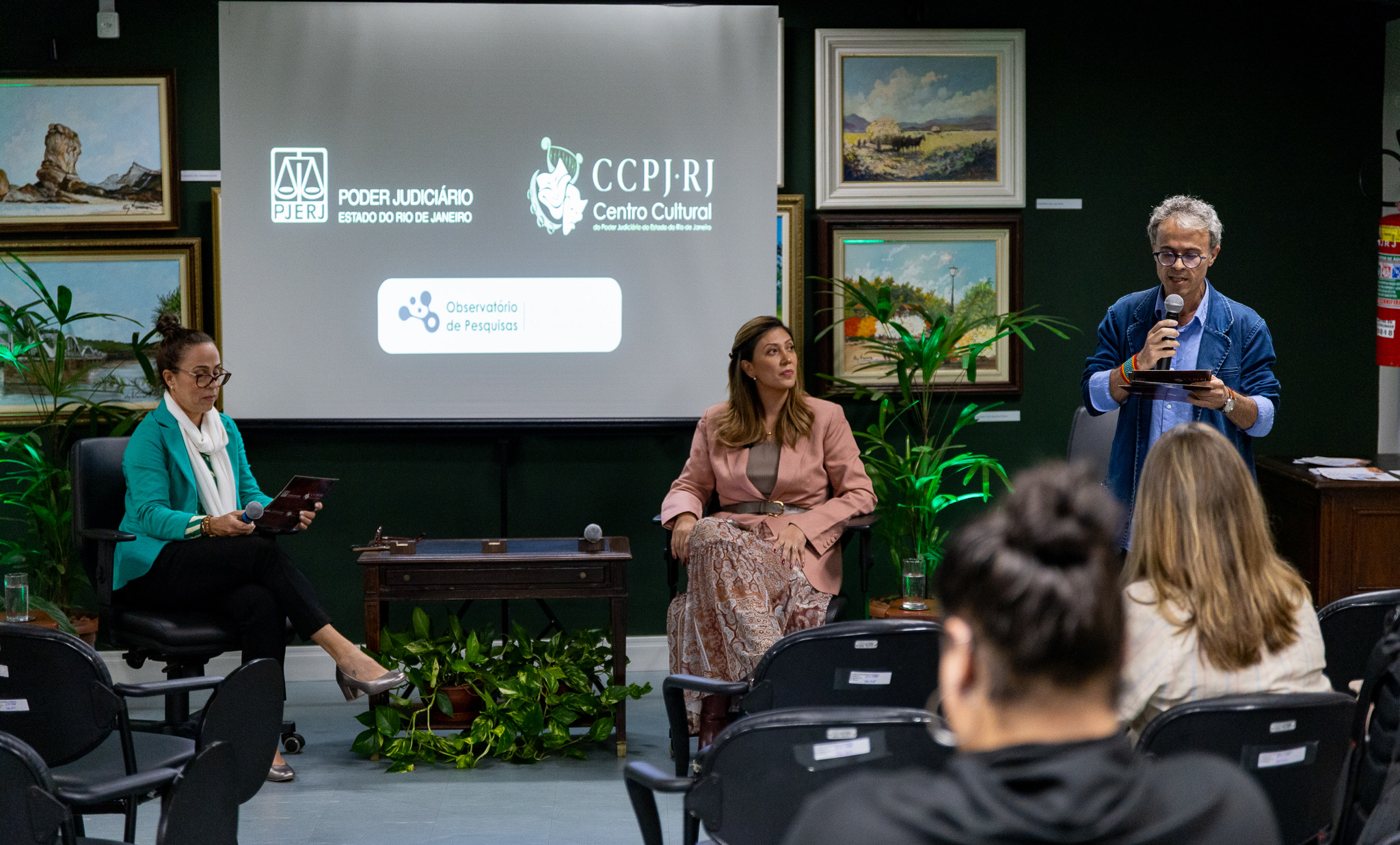 A imagem registra um evento realizado no Centro Cultural do Poder Judiciário do Estado do Rio de Janeiro (CCPJ-RJ). Em primeiro plano, um homem está em pé, segurando um microfone e falando ao público, enquanto lê anotações em um tablet. Ao seu lado, duas mulheres participam da mesa de debate, sentadas em poltronas, também com microfones disponíveis. Ao fundo, uma tela de projeção exibe as logomarcas do Poder Judiciário do Estado do Rio de Janeiro (PJERJ), do CCPJ-RJ e do Observatório de Pesquisas. O ambiente possui iluminação suave e decoração composta por plantas ornamentais e quadros com pinturas de paisagens distribuídos pelas paredes. Na plateia, algumas pessoas acompanham a apresentação sentadas em cadeiras voltadas para os palestrantes. 
