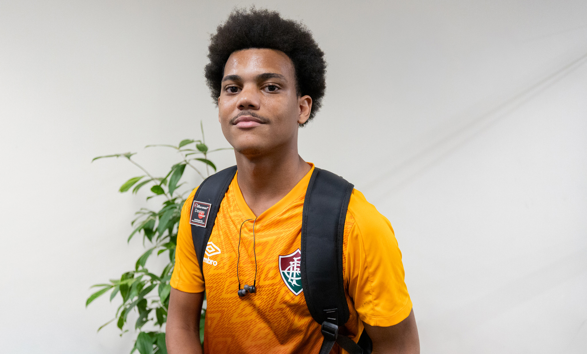 A imagem mostra um jovem em pé, posicionado de frente para a câmera, com expressão serena e um leve sorriso. Ele tem cabelos crespos volumosos e veste uma camisa esportiva laranja do Fluminense, além de uma mochila preta nas costas. Um par de fones de ouvido com fio está apoiado em seu pescoço. Ao fundo, há uma parede branca e uma planta ornamental de folhas verdes, compondo um ambiente interno simples e bem iluminado. A fotografia tem enquadramento em plano médio, com foco no retratado e fundo discretamente desfocado.