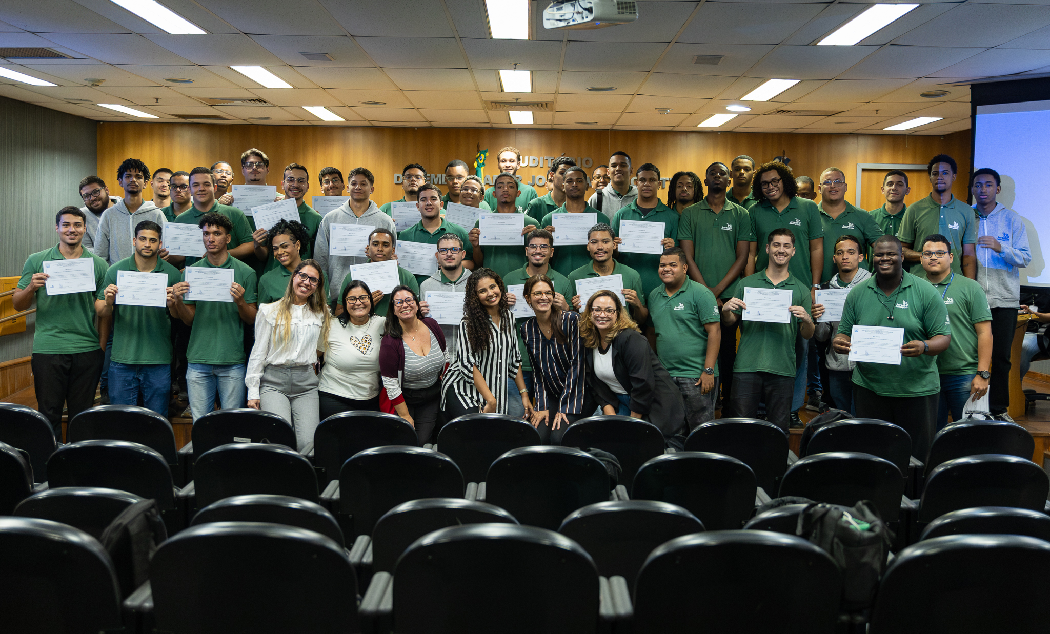 A imagem mostra um grande grupo de pessoas reunidas em um auditório durante uma cerimônia. A maioria dos participantes está vestida com camisas polo na cor verde e seguram certificados, exibindo-os para a foto. Na primeira fila, algumas mulheres posam sorrindo à frente do grupo. O ambiente possui paredes revestidas em madeira, iluminação de teto e uma tela de projeção ao lado direito. Ao fundo, é possível ver a identificação do auditório e bandeiras institucionais. Em primeiro plano aparecem diversas poltronas vazias do auditório, reforçando que a fotografia foi registrada a partir da área destinada ao público.
